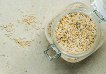 An open glass jar filled with uncooked brown rice, placed on a light textured surface with a few grains scattered around.