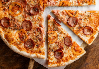 Thin-crust pizza topped with pepperoni, tomato slices, and herbs, partially sliced and served on parchment paper on a wooden surface.