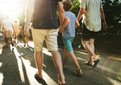 A group of people, including a child holding hands with two adults, walking down a sunlit path surrounded by trees.