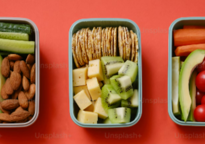 Three simple and light meal prep boxes with avocado, veggies, nuts, crackers, cheese, and fruit on a red background.