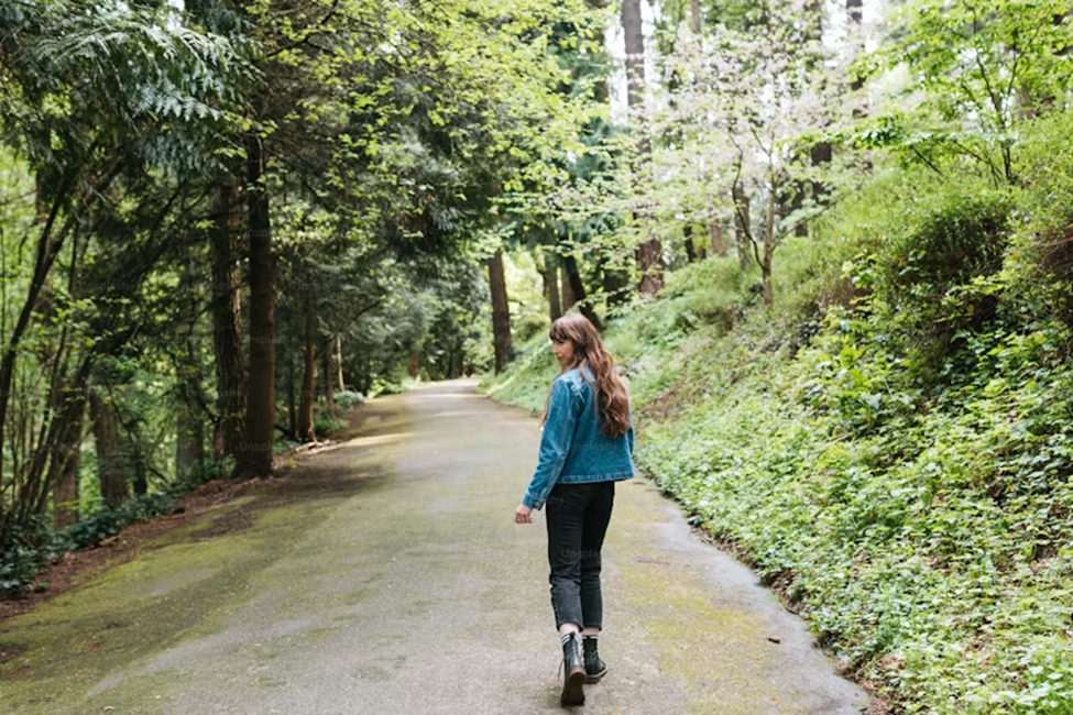 A woman with long hair wearing a denim jacket and boots walking down a shaded path surrounded by lush green trees and vegetation.