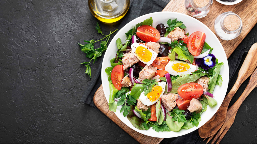 A fresh and vibrant salad in a white bowl featuring mixed greens, cherry tomatoes, boiled eggs, tuna chunks, red onion slices, and garnished with parsley, served on a wooden cutting board with a bottle of olive oil and wooden utensils nearby.