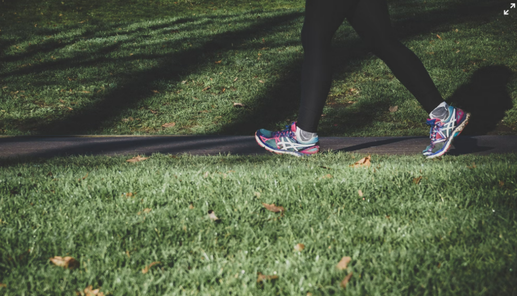 A person walking outdoors on a sunny day, wearing athletic shoes and leggings, surrounded by green grass and a peaceful park setting.