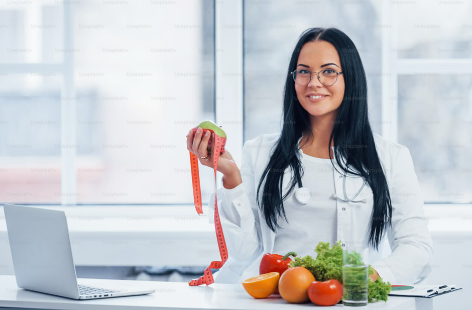 Smiling nutritionist holding a measuring tape and a green apple, surrounded by fresh fruits and vegetables, symbolizing a balanced approach to health and diet.