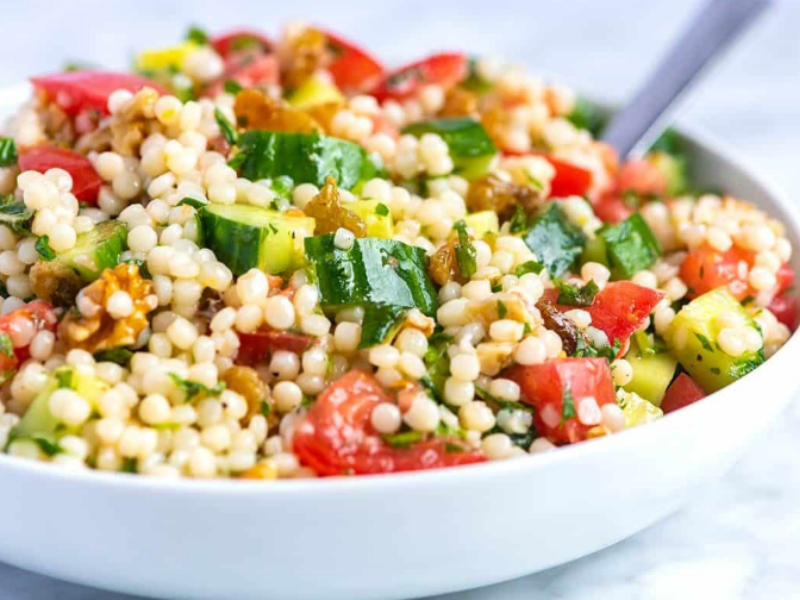 A bowl of pearl couscous salad with diced cucumbers, tomatoes, herbs, and golden raisins, served fresh.