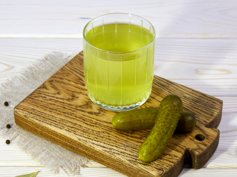 A glass of pickle juice and two small pickles on a wooden cutting board, placed on a white wooden surface with scattered bay leaves and peppercorns.