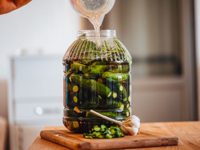 A large jar being filled with brine, containing cucumbers, garlic, and herbs, placed on a wooden cutting board with chopped green vegetables and a garlic bulb.