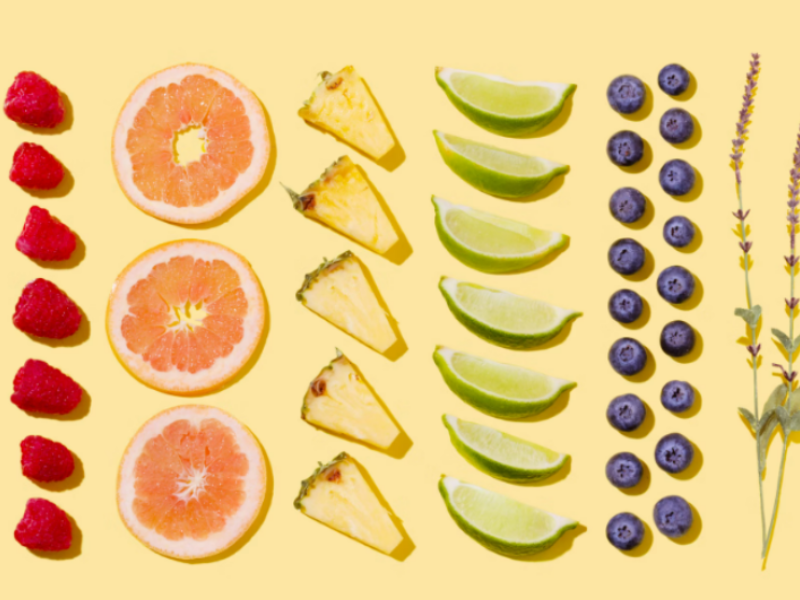 Colorful arrangement of fresh fruits and herbs on a yellow background, featuring raspberries, grapefruit slices, pineapple chunks, lime wedges, blueberries, and sprigs of lavender.