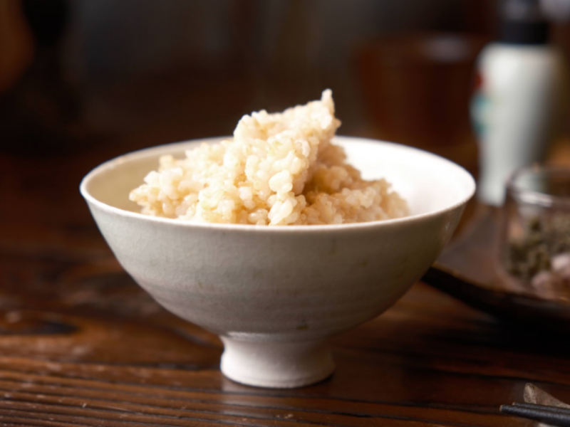 A bowl of freshly cooked brown rice served in a white ceramic bowl, placed on a wooden table with blurred background elements.